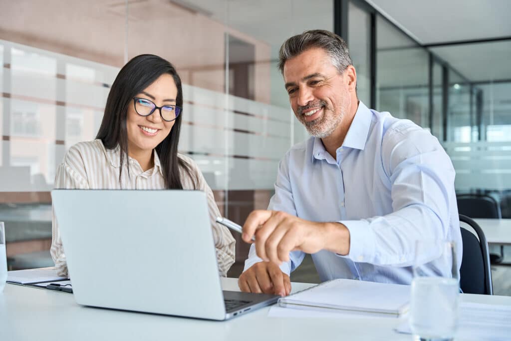 Mid aged Latin male manager mentor teaching young Asian female worker looking at laptop